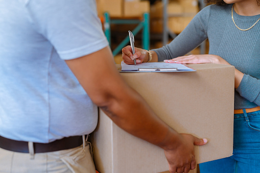 Writing a signature on order contract document paperwork for online shopping or postal package. Woman customer receiving a shipping, moving and shipment cardboard box from a delivery service worker