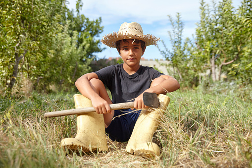 A kid is holding a hoe and he is helping with the farm work during his summer vacation