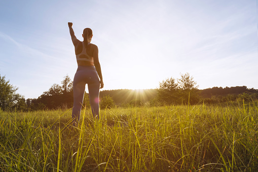 a girl in a fitness suit stands with her back to the camera at sunset with her hand raised up after a workout in the fresh air, motivational girl silhouette there is a place for an inscription