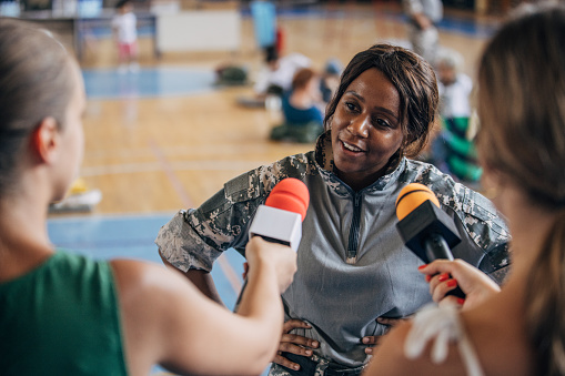 Diverse group of people, soldiers on humanitarian aid to civilians in school gymnasium, after natural disaster happened in city. Television press talking to a soldier.
