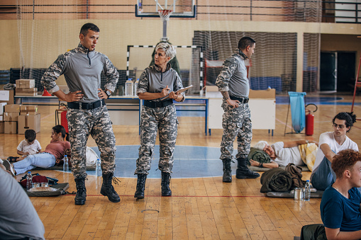 Diverse group of people, soldiers on humanitarian aid to civilians in school gymnasium, after natural disaster happened in city.