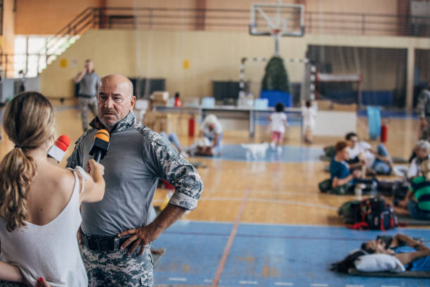 Diverse group of people, soldiers on humanitarian aid to civilians in school gymnasium, after natural disaster happened in city. Television press talking to a soldier.