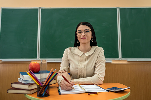 beautiful teacher with glasses sits at a desk with book, pencil notepad apple against blackboard, classroom