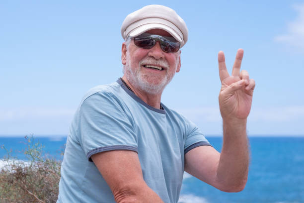 istock Smiling caucasian senior man with hat sitting at the beach looking at camera gesturing victory sign while enjoying vacation or retirement. Horizon over the water, copy space 1419571843