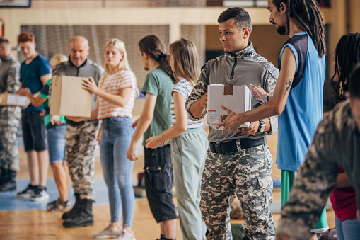 Diverse group of people, soldiers on humanitarian aid to civilians in school gymnasium, after natural disaster happened in city. Civilians assisting soldiers with donation boxes.