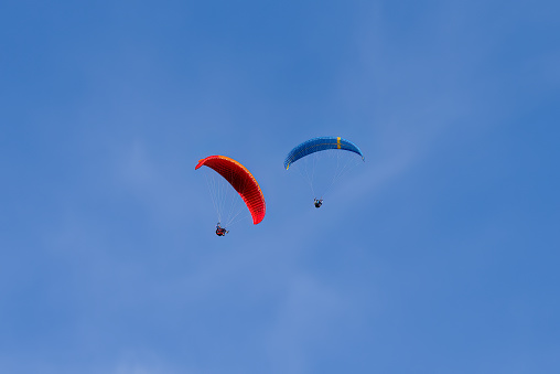 Pair of paragliders, one red and orange and the other blue that fly in the sky together.