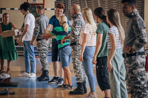 Diverse group of people, soldiers on humanitarian aid to civilians in school gymnasium, after natural disaster happened in city. Civilians assisting soldiers with donation boxes.