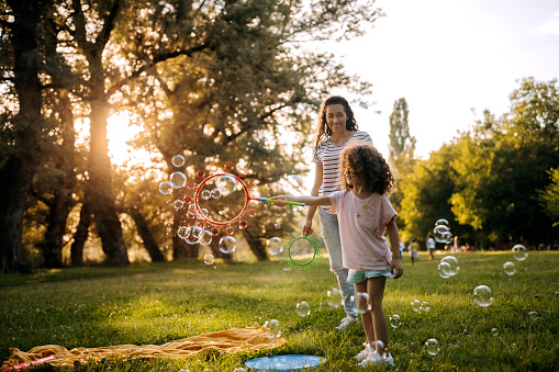 Cute little girl playing with bubble wand toy in a public park with her mother