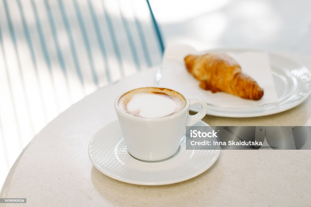 Cup of cappuccino and a croissant on a table in a street cafe. Selective focus. Cup of cappuccino and a croissant on a table in a street cafe. Selective focus. Place for text. Coffee - Drink Stock Photo