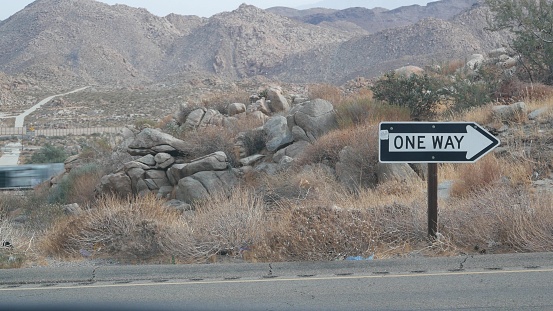 One way road transport sign with arrow on roadside or wayside, highway or freeway in California USA. Road trip in desert valley among mountains and hills with boulders. Western wilderness nature.