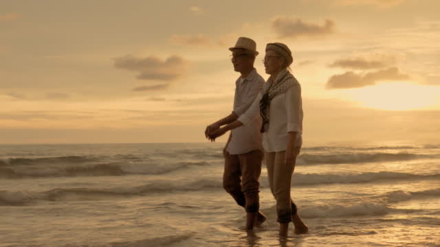 Asian Senior couple talking and walking along to sunset sea surf on sand beach.