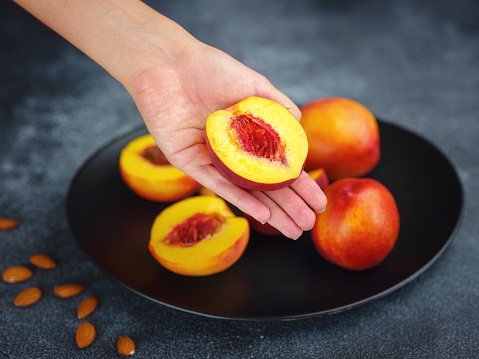 Hand holding fresh peach fruit and black dish with peaches and cloth on table blurred background. Top view. Close-up photo. Healthy food and fruit concept