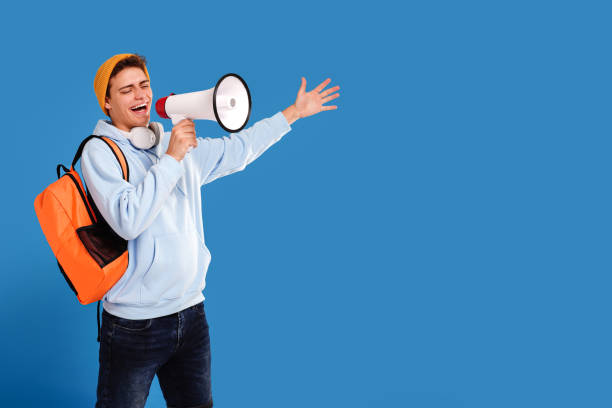 jovem feliz em roupas da moda e mochila posando isolado no fundo do estúdio azul, gritando algumas boas notícias no megafone. educação, conceito de estilo de vida estudantil. - university education screaming shouting - fotografias e filmes do acervo