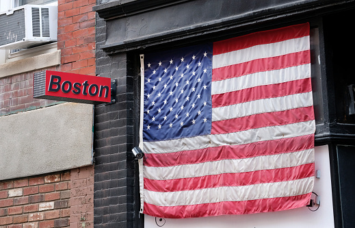 A sign with the word Boston next to an American flag hanging over a window