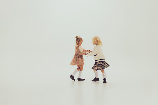Portrait of little beautiful girls, children holding hands and spinning, playing isolated over grey studio background. Concept of childhood, friendship, fun, lifestyle, fashion, retro style