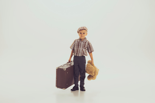 Portrait of little boy, child in checkered shirt posing with vintage suitcase and bear toy isolated over grey studio background. Concept of childhood, friendship, fun, lifestyle, fashion, retro style