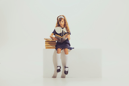 Portrait of beautiful school girl, sitting in school uniform and reading book isolated over grey studio background. Concept of childhood, friendship, fun, lifestyle, fashion, retro style