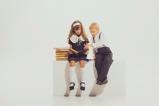 Portrait of school girl and boy, sitting in uniform and reading book isolated over grey studio background. Homework. Concept of childhood, friendship, fun, lifestyle, fashion, retro style