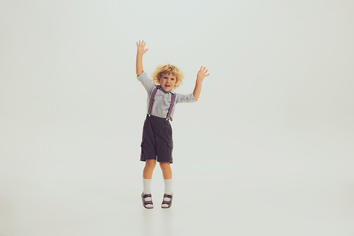 Portrait of little boy, child in suspenders and shorts playing, posing, having fun isolated over grey studio background. Concept of childhood, friendship, fun, lifestyle, fashion, retro style
