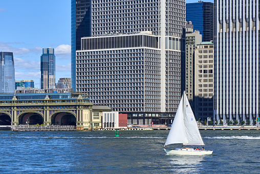 New York, NY - August 13, 2022: A small sailboat on the East River in front of Lower Manhattan skyscrapers on a sunny summer day. The Battery Maritime Building and ferry terminal are behind.