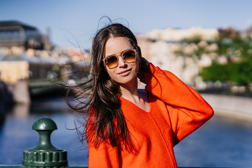 Portrait of brunette female model posing outdoors in glasses, red sweater walking against blurry old town. Awesome brunette Italian woman with loose fluttering hair traveling. Beautiful people.