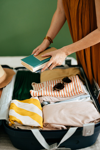 Anonymous woman in  an orange dress holding books ready to put them in an open suitcase on the bed. There are already clothes in the suitcase, a hat and sunglasses.