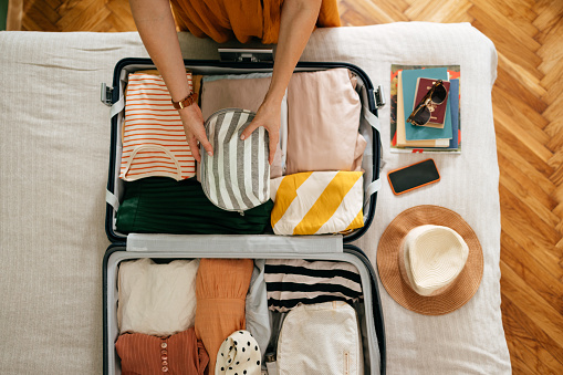 Shot from above of an anonymous woman packing things in her suitcase on the bed. She is holding and putting a grey and white stripped cosmetics bag in. There is her mobile phone, books and sunglasses on the bed.