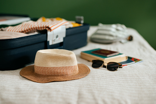 Close up shot of an open suitcase with various stuff laid on the bed ready for a summer vacation trip away. There is a brown hat, sunglasses, books and a passport. There is no one on the photo.