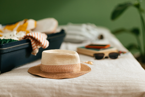 Close up shot of an open suitcase with various stuff laid on the bed ready for a summer vacation trip away. There is a brown hat, sunglasses, books and a passport. There is no one on the photo.