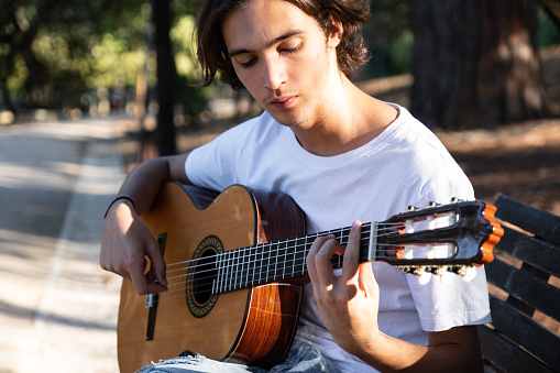 Guitarist plays on guitar outdoors.  Handsome guy plays the guitar on the nature sitting on a bench.  Young man plays a musical instrument outside  house. Musician plays a classical guitar in the park.