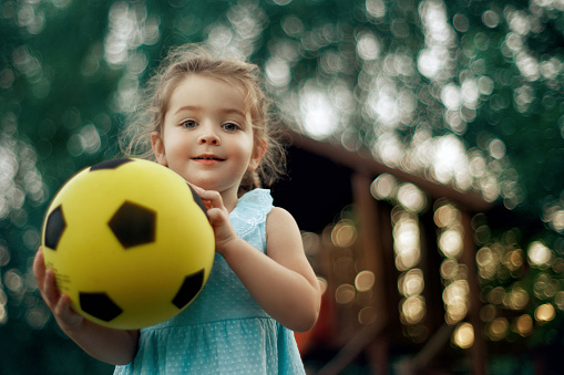 Close up portrait with cute little girl wearing a dress , holding a yellow soccer ball in her hand , she is in the park