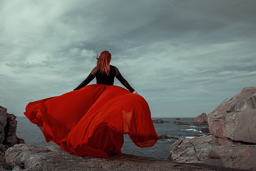 Redhead Woman in long red dress standing on the rock at coastline admiring the view . Scenic view