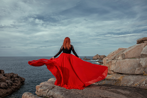 Fashion concept of young woman in red flying dress with red hair on a rock at coastline admiring the ocean