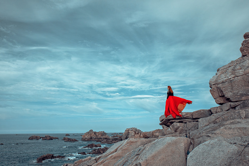 Side view of red head woman wearing long red dress at the coastline, looking at the sea view