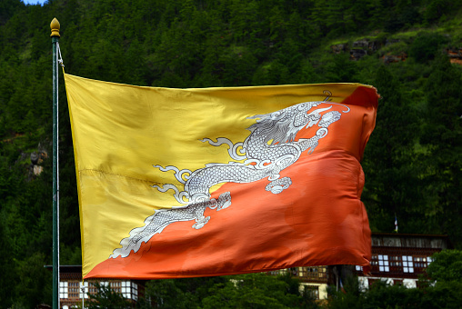 Flag of Bhutan in the wind - The two colors of the flag, divided diagonally, represent spiritual and temporal power within Bhutan. The orange part of the flag represents the Drukpas monasteries and Buddhist religious practice, while the saffron yellow field denotes the secular authority of the dynasty. Regarding the dragon, it represents Druk, the Tibetan name for the kingdom of Bhutan. The jewels clamped in the dragon's claws symbolize wealth. The snarling mouth represents the strength of the male and female deities protecting the country.