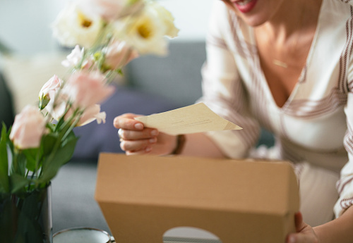 Close up shot of an anonymous woman sitting and in her hands she is holding a paper note. There is an opened carton gift box on the coffee table in front of her and a vase with flowers.