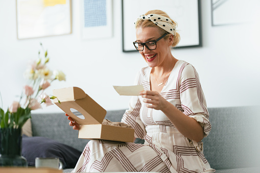 Beautiful cheerful woman in a dress, sitting on a sofa at home and opening a gift box that has arrived for her. She is excited while holding and reading a paper that she got. There is an opened carton gift box on the coffee table in front of her.