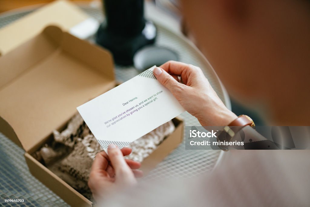 Anonymous Woman Reading a Note From a Gift Box (Copy Space) Anonymous woman sitting and in her hands  holding a paper note with a written message on it. There is an opened carton gift box on the coffee table in front of her and a cup of coffee. Unboxing Stock Photo