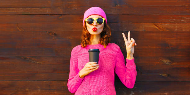 Portrait of stylish modern young woman blowing her lips sends sweet air kiss with cup of coffee wearing colorful pink hat, knitted sweater on brown wooden wall background stock photo