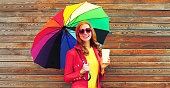 Autumn portrait of happy smiling young woman with colorful umbrella and cup of coffee wearing red jacket, heart shaped sunglasses on city street