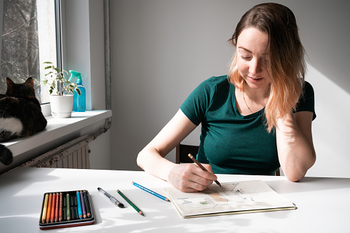 Young woman in green t shirt sitting at the table by the window and drawing with paper and pencils. Cat lying on window sill.