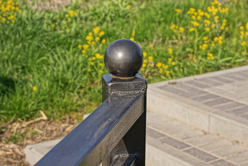 part of a black iron handrail with a ball near the gray concrete steps and green grass with yellow flowers in the park