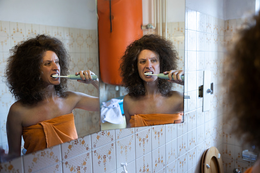 Mid Adult Curly hair Woman Brushing Her Teeth with an Electric Brush in Front of Domestic Bathroom Mirror