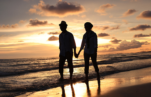 Senior couple holding hands for support walk on beach , love and care with outdoor wellness, retirement and holiday lifestyle with sunset sky