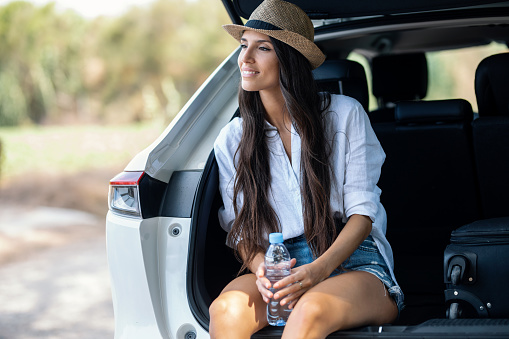 Shot of beutiful woman sitting in the trunk of the car while drinking water in the forest.
