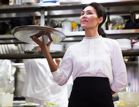 Confident positive waitress holding delicious cooked meals in restaurant kitchen