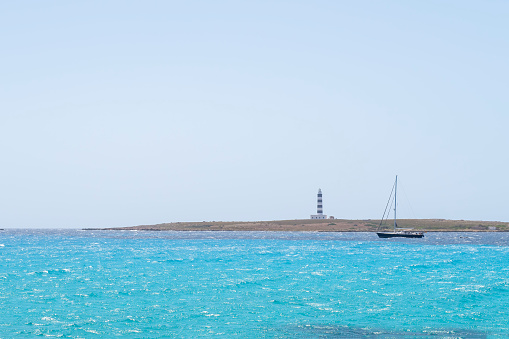 a sailboat skirts a cape with a blue and white lighthouse, on a sea of turquoise waters, clear sky, Isla del Aire, Punta Prima, Menorca, Spain, copy space