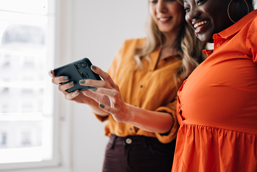 A beautiful smiling Caucasian influencer watching something on her smartphone with her charming female friend.