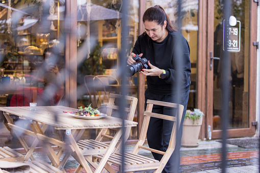 A woman, a photographer, is taking food photos for a blog on a table of an outdoor restaurant using a professional camera.