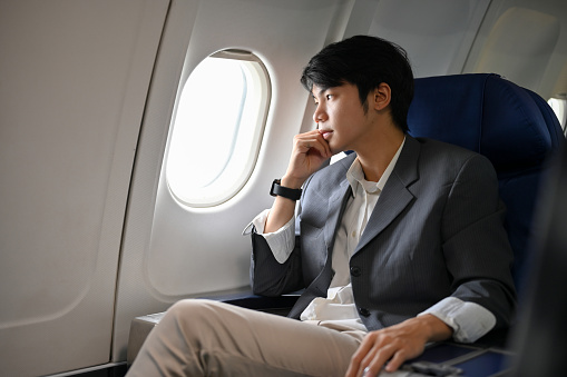 Handsome and smart young Asian businessman or business boss in formal suit sits at the window seat in a business class, looking out the window, pensively thinking and planning his business project.